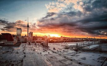 A view of Berlin at sunset, featuring city buildings with the TV Tower prominently visible under a dramatic sky.
