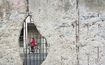 A person cycling past a hole in a section of the Berlin Wall, seen through a wire barrier.