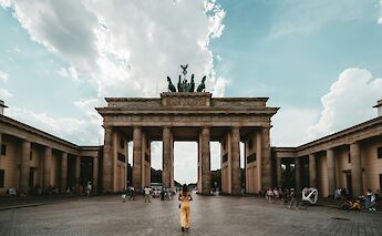 A person standing in front of the Brandenburg Gate in Berlin, with a partly cloudy sky above.