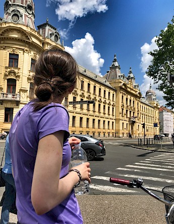 A person holding a water bottle, standing near an ornate historic building in Zagreb, Croatia. The building features decorative architecture, with traffic and pedestrians in the vicinity.