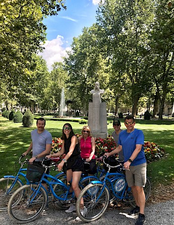 A group of people posing with bicycles in a park in Zagreb, Croatia. They are standing near a flower bed with a bust statue and a fountain in the background, surrounded by trees.