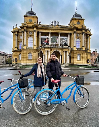 Two people stand with bicycles in front of a grand historic building with a yellow facade and arched entrance in Zagreb, Croatia.