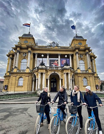 Four people with bicycles are gathered in front of a historic building with a decorative facade and flags at the top in Zagreb, Croatia.
