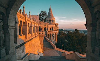 An architectural view of Buda Castle's neo-Gothic style, bathed in golden sunset light, with arches, towers, and distant city views.