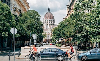 Cyclists ride through a tree-lined street in Budapest, with a large domed building visible in the background.