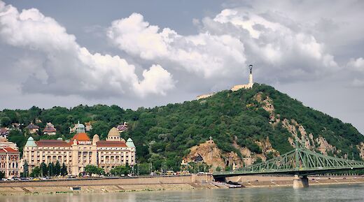 Gellert Hill rises above the Danube River in Budapest, featuring lush greenery and a prominent hotel at the base.