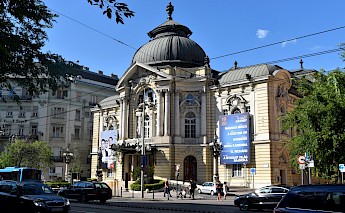 A grand building with domes and ornate details, identified as the Operetta Theatre in Budapest, is surrounded by street activity and trees.