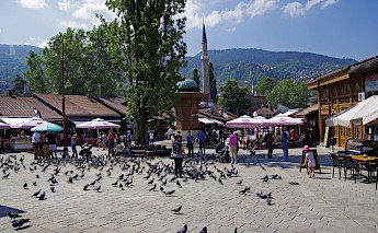 People and pigeons at Bascarsija square in Sarajevo, with a minaret and surrounding buildings.