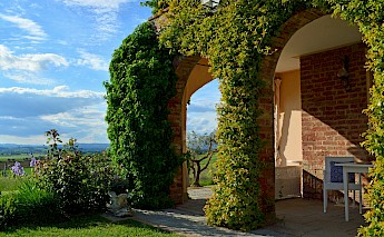 A villa with ivy-covered arches overlooks a scenic countryside with a blue sky in Tuscany.