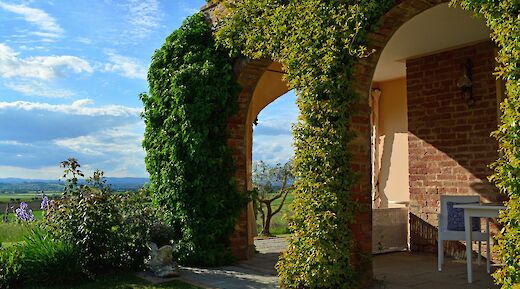 A villa with ivy-covered arches overlooks a scenic countryside with a blue sky in Tuscany.