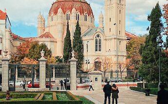 A grand building with a domed roof and tall tower, surrounded by trees and people walking along a&hellip;
