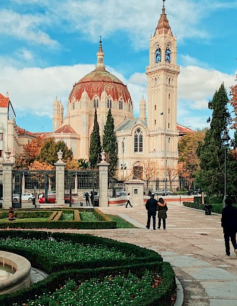 A grand building with a domed roof and tall tower, surrounded by trees and people walking along a path in Madrid, Spain.