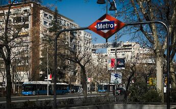 A metro entrance sign for Santiago Bernabeu station with city buildings and trees in the background.
