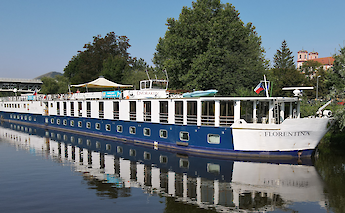 A large blue and white vessel named Florentina docked alongside a river, with trees and a bridge in…