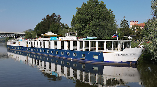 A large blue and white vessel named Florentina docked alongside a river, with trees and a bridge in the background.