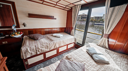 A cozy cabin interior with two beds, wooden walls, a bowl of fruit, and large windows overlooking a river.