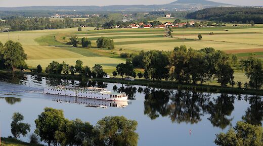 A white vessel gliding along a calm river, surrounded by green fields and trees, with a hill in the distant background.