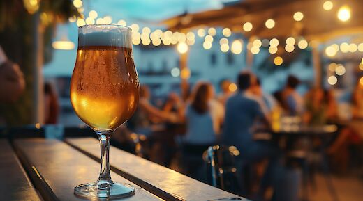 A glass of beer on a table in an outdoor setting with blurred lights and people in the background.