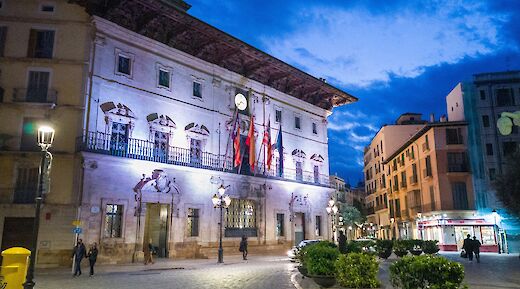A historic building in Plaza de Cort, Palma de Mallorca, illuminated at night with people walking nearby.