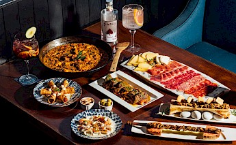 An assortment of Spanish tapas, including cured meats, fried rolls, paella, and olives, arranged on a wooden table with drinks.