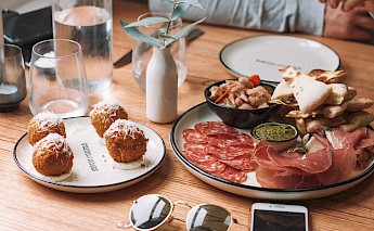 A variety of tapas on a wooden table, including meats, bread, and cheeses, with glasses, a phone, and sunglasses nearby.
