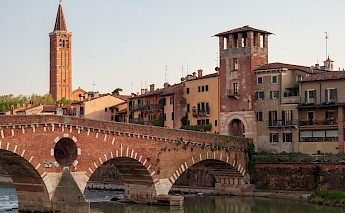 A historic brick bridge arches over a river in Verona, Italy, with colorful buildings and a tall bell tower in the background.