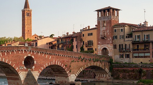 A historic brick bridge arches over a river in Verona, Italy, with colorful buildings and a tall bell tower in the background.