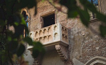 A historic balcony with Gothic arches on a brick building in Verona, associated with the Romeo and Juliet story, partially obscured by leaves.