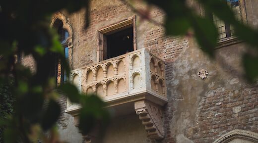 A historic balcony with Gothic arches on a brick building in Verona, associated with the Romeo and Juliet story, partially obscured by leaves.