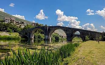 A stone bridge with multiple arches spans a tranquil river in Trebinje, Bosnia and Herzegovina, surrounded by greenery and hills.
