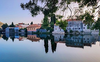 A calm river reflects colorful and rustic buildings in Trebinje, Bosnia and Herzegovina, with tree branches framing the scene.