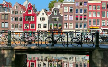 Bicycles are parked on a bridge over a canal in Amsterdam, with colorful buildings in the background reflecting in the water.