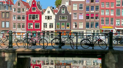 Bicycles are parked on a bridge over a canal in Amsterdam, with colorful buildings in the background reflecting in the water.