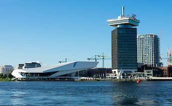 A view of modern buildings by the waterside in Amsterdam, Holland, with cranes in the background under a clear blue sky.