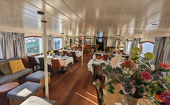 A dining and lounge area inside a boat, featuring tables set for a meal, comfortable seating, and…