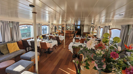 A dining and lounge area inside a boat, featuring tables set for a meal, comfortable seating, and large windows with curtains. A vase with assorted flowers is placed on a table.