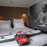 A cozy bedroom with twin beds, white linens, and an artistic mural of a woman pouring milk, possibly inspired by Vermeer's painting. Two tulips and a red bag with "Boat Bike Tours" are placed on the bed.