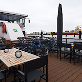 The deck of a boat set up with wooden tables and chairs branded "Magnifique I," surrounded by numerous bicycles against a harbor backdrop.