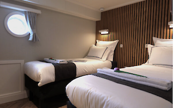 A cabin with two single beds, finished with white bedding and dark accents. A small circular window…