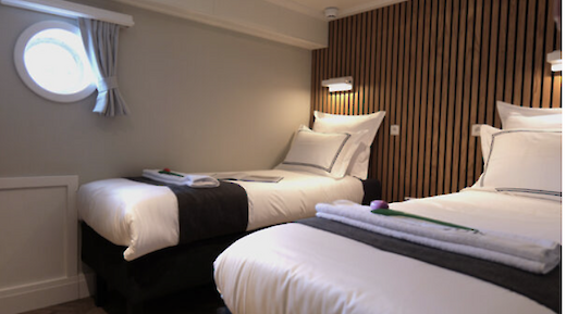 A cabin with two single beds, finished with white bedding and dark accents. A small circular window offers a glimpse outside, and the walls are adorned with wood paneling.