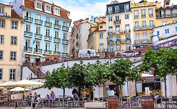 A lively public square in Lisbon with colorful buildings and outdoor café seating under trees.