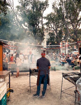 A bustling outdoor food market in Lisbon with people gathered around tables while a vendor grills food.