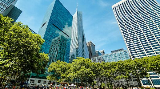 Tall skyscrapers with reflective glass facades rise above lush trees in New York City under a clear blue sky.