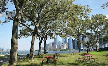 A park with red picnic tables and trees in the foreground, with the New York City skyline and Hudson River visible in the background.