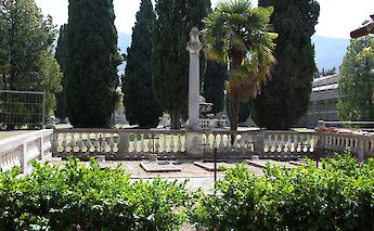 The garden area with a central stone column and railings, surrounded by tall cypress trees, located in Calci, Pisa, Italy.