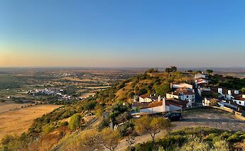 A scenic view of the Alentejo region in Portugal, showing a hillside village with white buildings and red roofs, surrounded by expansive fields under a clear blue sky.