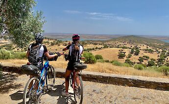 Two cyclists with mountain bikes are standing on a stone path, overlooking a vast rural landscape with rolling hills and a distant reservoir under a clear blue sky in Portugal.