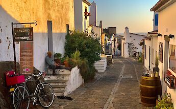 A picturesque cobblestone street in a Portuguese village during sunset, with a person sitting on steps next to a wall and a bicycle leaning against it.
