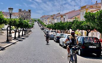Cyclists riding through a cobblestone street flanked by trees and parked cars in a Portuguese town, with a historic building visible at the end of the road.