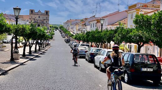 Cyclists riding through a cobblestone street flanked by trees and parked cars in a Portuguese town, with a historic building visible at the end of the road.